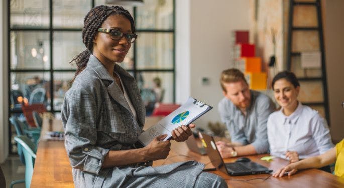 A woman sitting on a table where her colleagues are gleefully planning work. She's holding a clipboard with a pie chart, triumphantly looking at the camera.