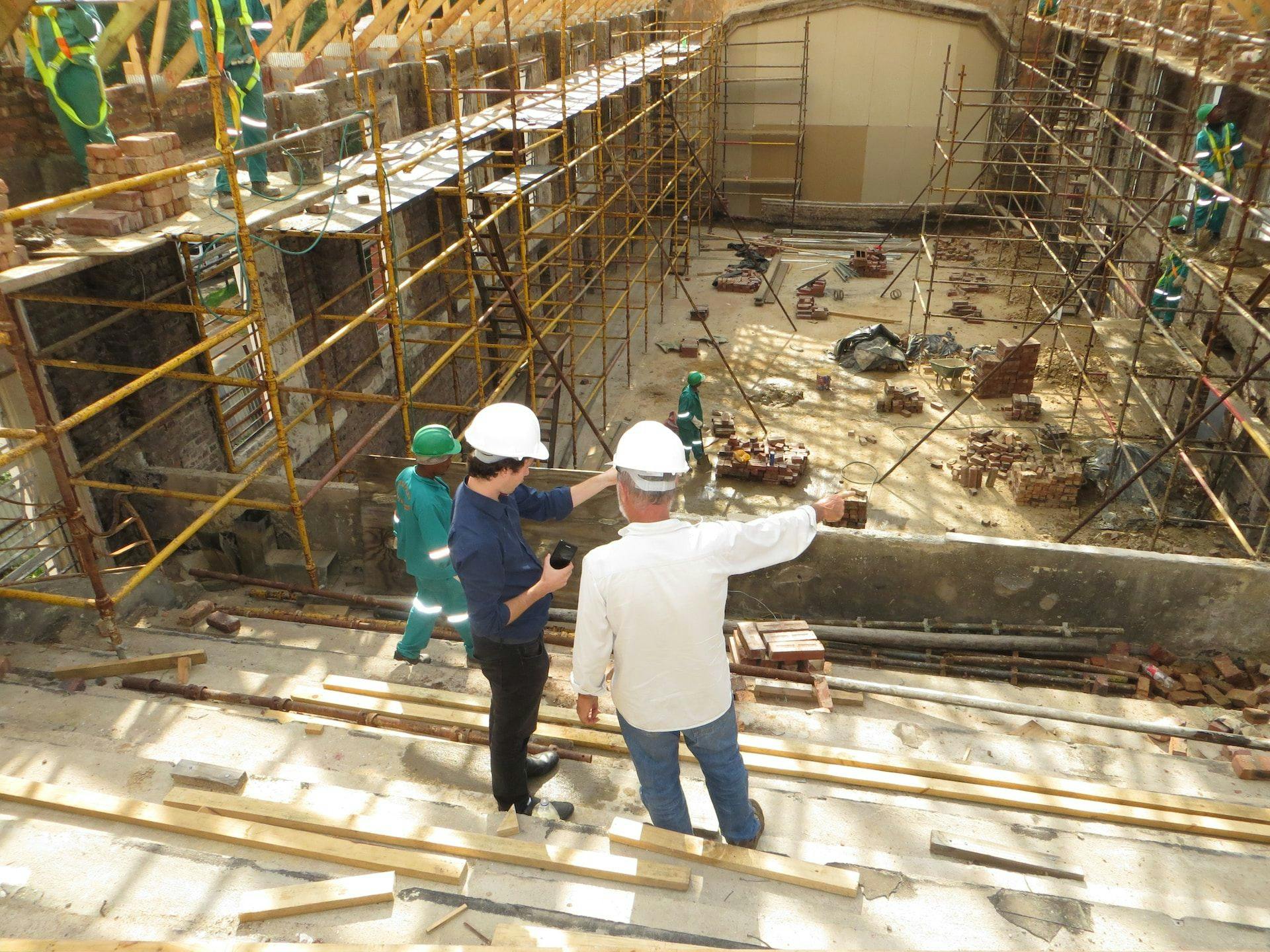 Construction workers wearing hard hats and reflective vests inside a vast building that is currently being constructed.