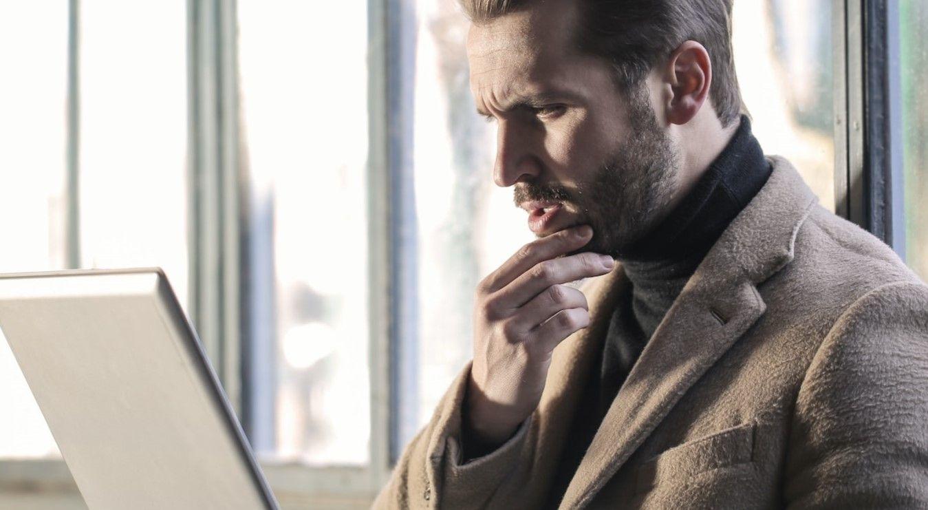 A gentleman wearing a coat is seated on a bench while working on his laptop.