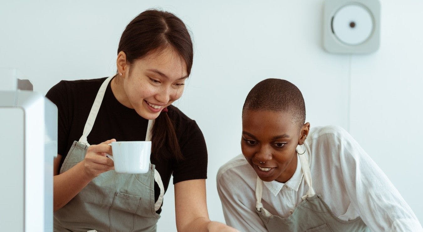 Two female individuals wearing aprons are collaborating on a laptop computer.