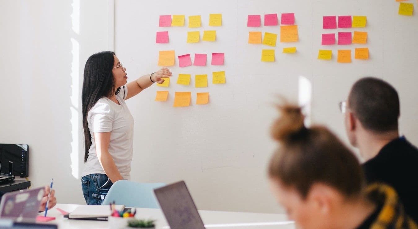 A female individual is positioned in front of a whiteboard that is adorned with adhesive notes.